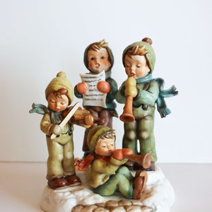 K&ouml;nnte beinhalten: Eine Figuren-Gruppe aus vier Kindern in Winterkleidung, die Musik machen. Die Kinder tragen blaue, gr&uuml;ne und braune Kleidung. Die Kinder spielen Violine, Trompete, Blockfl&ouml;te und singen von einem Notenblatt. Die Figuren-Gruppe steht auf einem wei&szlig;en Sockel mit einer braunen, schneebedeckten Oberfl&auml;che.