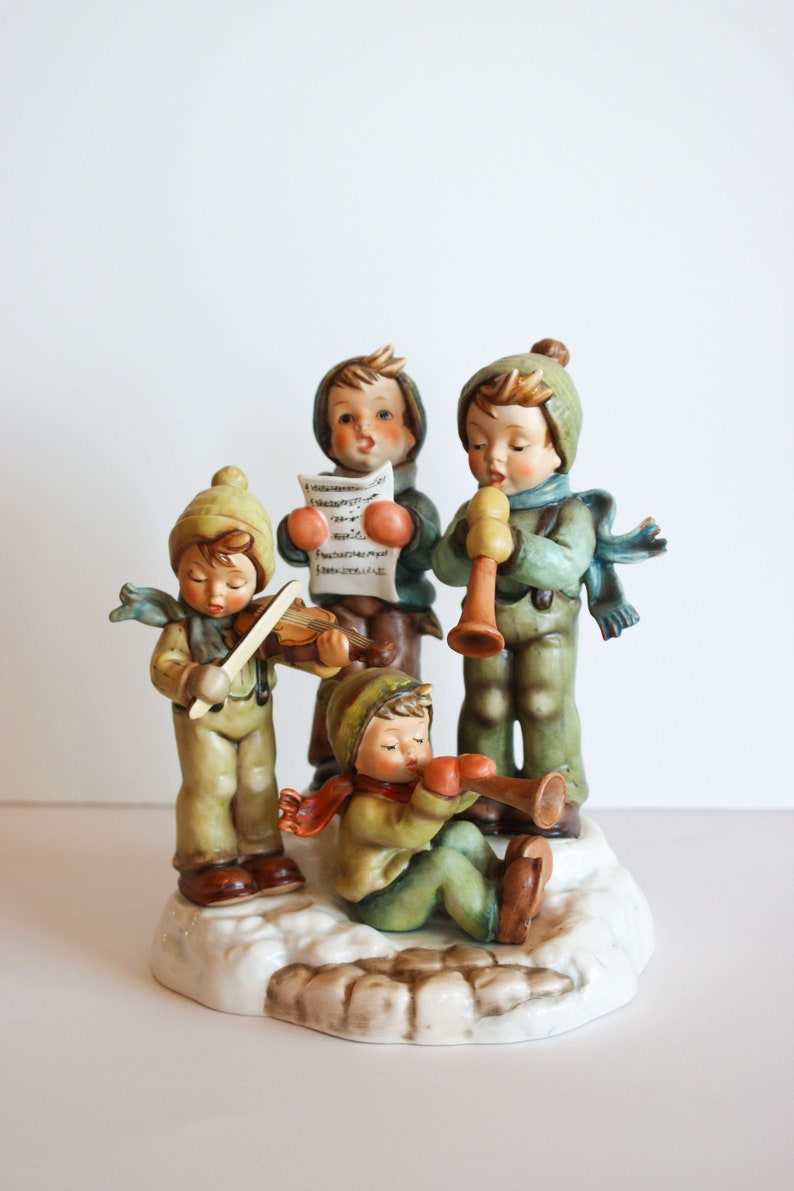 K&ouml;nnte beinhalten: Eine Figuren-Gruppe aus vier Kindern in Winterkleidung, die Musik machen. Die Kinder tragen blaue, gr&uuml;ne und braune Kleidung. Die Kinder spielen Violine, Trompete, Blockfl&ouml;te und singen von einem Notenblatt. Die Figuren-Gruppe steht auf einem wei&szlig;en Sockel mit einer braunen, schneebedeckten Oberfl&auml;che.