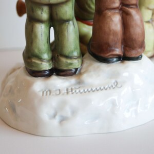 K&ouml;nnte beinhalten: Eine Porzellanfigur von drei Kindern, die auf einem schneebedeckten Sockel stehen. Die Figur ist signiert mit "M.J. Hummel" und "Goebel".
