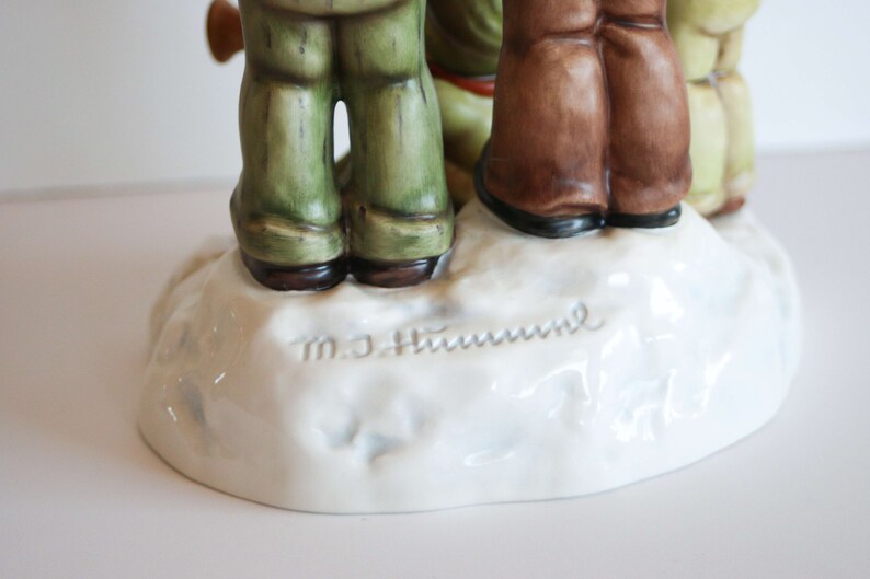 K&ouml;nnte beinhalten: Eine Porzellanfigur von drei Kindern, die auf einem schneebedeckten Sockel stehen. Die Figur ist signiert mit "M.J. Hummel" und "Goebel".