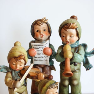 K&ouml;nnte beinhalten: Eine Gruppe von vier Porzellanfiguren von Kindern, die in Winterkleidung gekleidet sind. Die Kinder singen Weihnachtslieder, spielen Violine und Fl&ouml;te. Die Figuren sind realistisch detailliert bemalt und haben einen Vintage-Look.