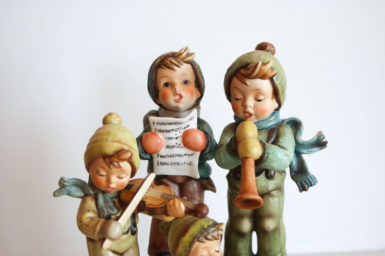 K&ouml;nnte beinhalten: Eine Gruppe von vier Porzellanfiguren von Kindern, die in Winterkleidung gekleidet sind. Die Kinder singen Weihnachtslieder, spielen Violine und Fl&ouml;te. Die Figuren sind realistisch detailliert bemalt und haben einen Vintage-Look.