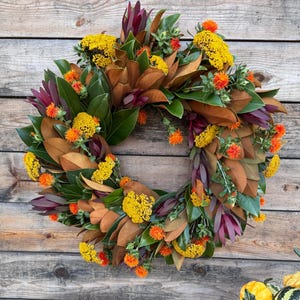 May include: A circular wreath featuring deep green and brown magnolia leaves, accented with clusters of bright yellow yarrow flowers and vibrant orange safflower blooms, arranged in a dense, layered pattern for a seasonal decorative display.