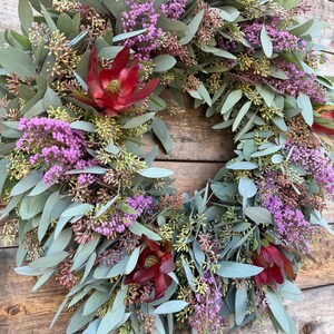May include: A circular wreath composed of eucalyptus leaves, purple flowers, and deep red blooms. The wreath is set against a rustic wooden background, showcasing the natural textures and colors of the floral arrangement. The design is full and lush.