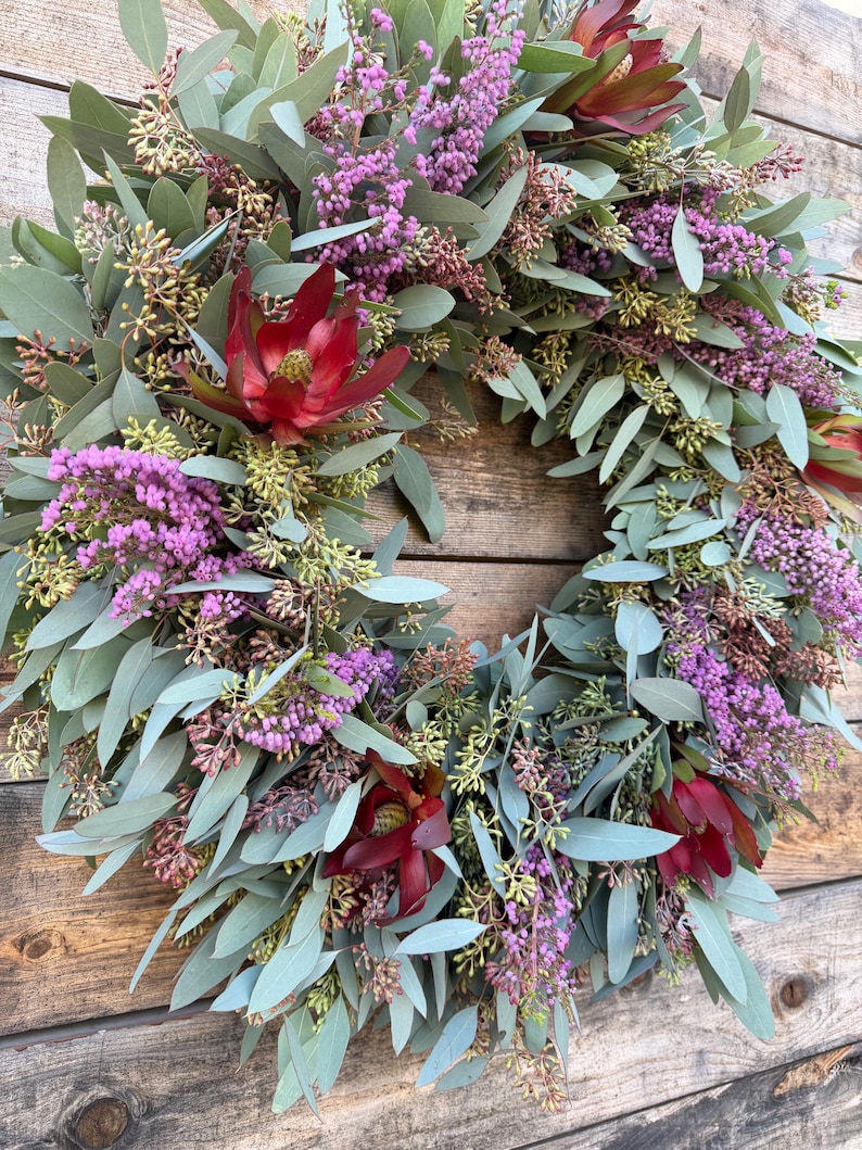 May include: A circular wreath composed of eucalyptus leaves, purple flowers, and deep red blooms. The wreath is set against a rustic wooden background, showcasing the natural textures and colors of the floral arrangement. The design is full and lush.