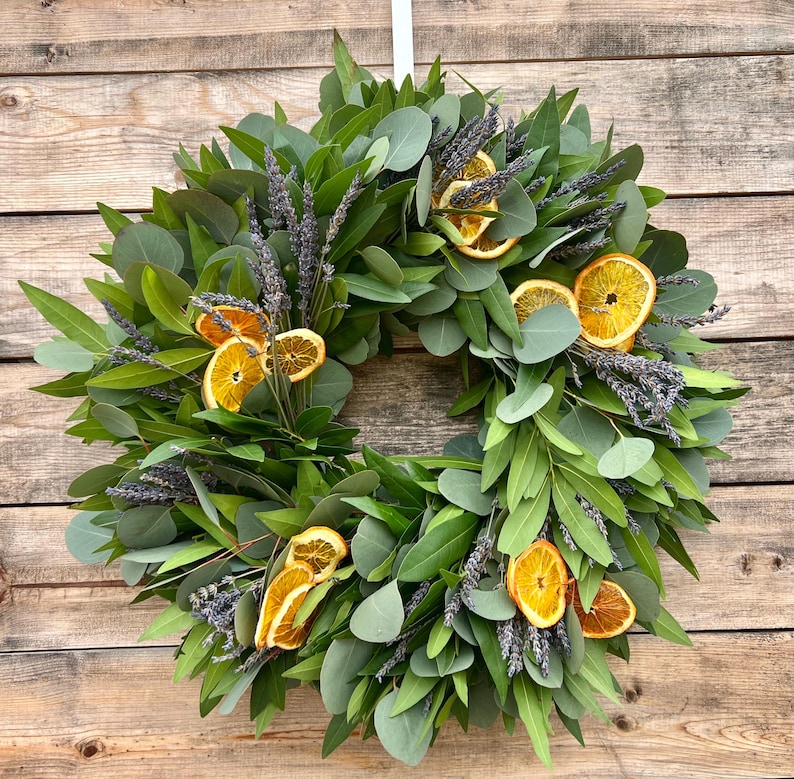 May include: A circular wreath composed of green eucalyptus leaves, sprigs of lavender, and dried orange slices. The wreath is displayed against a wooden background, creating a natural and rustic aesthetic. The arrangement is suitable for home decor.