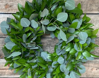 May include: A fresh, green wreath composed of various types of foliage. The wreath features a mix of bright green and silvery-green leaves, creating a natural and textured appearance. It is suspended against a wooden background.