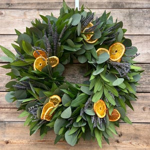 May include: A circular wreath featuring green eucalyptus leaves, sprigs of lavender, and dried orange slices. The wreath is suspended against a wooden background. The natural elements create a rustic and inviting aesthetic.