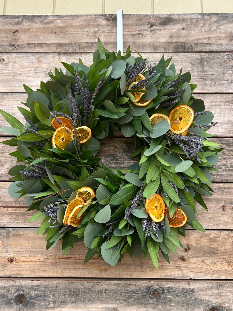 May include: A circular wreath featuring green eucalyptus leaves, sprigs of lavender, and dried orange slices. The wreath is suspended against a wooden background. The natural elements create a rustic and inviting aesthetic.