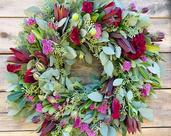 Op de afbeelding: Een levendige bloemenkrans met een mix van groen, bordeauxrode en paarse bloemen. De krans bestaat uit eucalyptusbladeren, bordeauxrode protea's en trossen paarse bloemen. Het wordt getoond tegen een houten achtergrond.