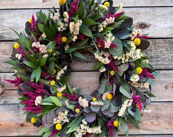 Può includere: Una ghirlanda circolare composta da vari fiori e fogliame secchi. La ghirlanda presenta foglie verdi, foglie viola scuro e grappoli di piccoli fiori bianchi, gialli e magenta. Su sfondo di legno.