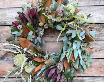 May include: A circular wreath featuring deep burgundy protea flowers, green eucalyptus leaves, brown magnolia leaves, and pale green banksia blooms. The arrangement includes wispy dried grasses and small filler foliage, creating a textured botanical display.