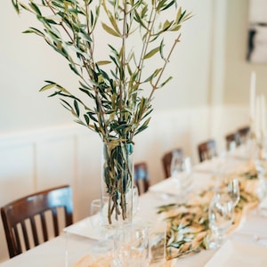 May include: A clear glass vase filled with olive branches sits on a table set for a formal event. The table is covered with a white tablecloth and decorated with a gold runner and glassware.