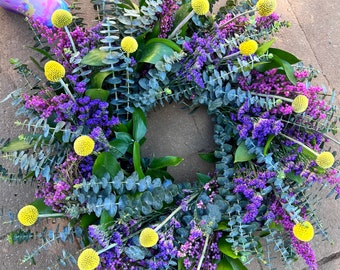 May include: A wreath made of eucalyptus, purple and pink flowers, and yellow pom poms.