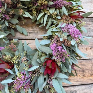 May include: A circular wreath composed of eucalyptus leaves, purple flowers, and burgundy blooms. The wreath is arranged on a wooden surface, showcasing a natural and rustic aesthetic. The leaves are a muted green, and the flowers add pops of color.
