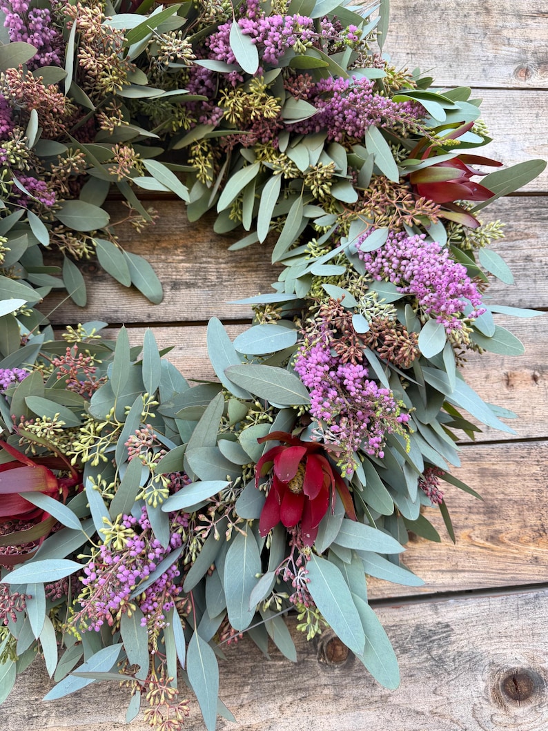 May include: A circular wreath composed of eucalyptus leaves, purple flowers, and burgundy blooms. The wreath is arranged on a wooden surface, showcasing a natural and rustic aesthetic. The leaves are a muted green, and the flowers add pops of color.