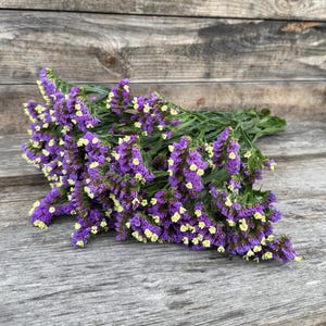 May include: A bundle of purple and yellow statice flowers, with green stems, rests on a weathered wooden surface. The flowers have small, clustered blooms and are arranged in a natural, organic style. The background is a rustic wooden panel.