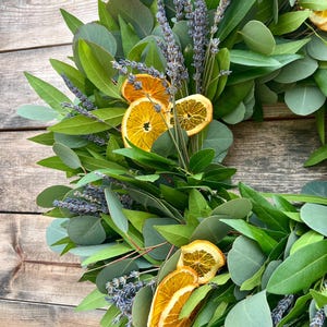 May include: A handmade wreath featuring green eucalyptus leaves, sprigs of lavender, and dried orange slices. The wreath is arranged in a circular shape, with the natural elements creating a rustic and aromatic display. The background is a wooden surface.