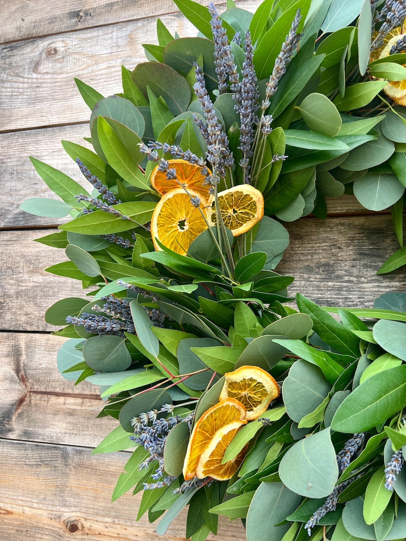 May include: A handmade wreath featuring green eucalyptus leaves, sprigs of lavender, and dried orange slices. The wreath is arranged in a circular shape, with the natural elements creating a rustic and aromatic display. The background is a wooden surface.