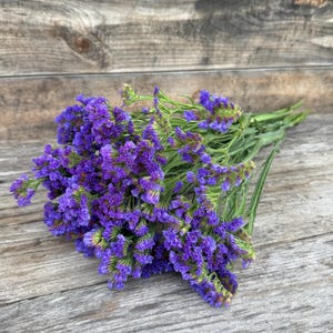 May include: A bouquet of purple statice flowers with green stems and leaves. The flowers are a vibrant shade of purple, with a textured appearance. The bouquet is arranged on a weathered wooden surface.