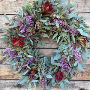 May include: A circular wreath composed of green eucalyptus leaves, purple flowers, and deep red protea blooms. The wreath is displayed against a rustic wooden background, creating a natural and inviting aesthetic. Perfect for home decor.