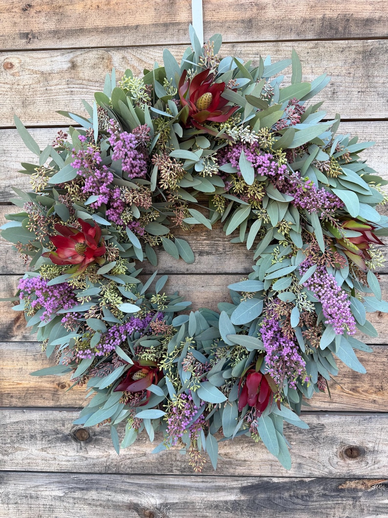 May include: A circular wreath composed of green eucalyptus leaves, purple flowers, and deep red protea blooms. The wreath is displayed against a rustic wooden background, creating a natural and inviting aesthetic. Perfect for home decor.