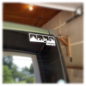 May include: A black car with a white sticker on the back window. The sticker has four images of a city skyline, a car driving on a road, a forest, and a mountain range. The text "element" is below the images.