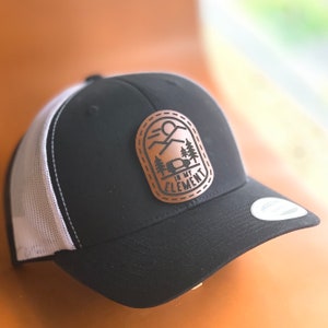 May include: Black and white trucker hat with a brown leather patch that says "In My Element" with a mountain and camper van design.