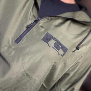 May include: Olive green anorak jacket with a black zipper and hood drawstring. The jacket features a black rectangular patch with a topographic design and a vehicle silhouette. The jacket is made of a lightweight, water-resistant material.