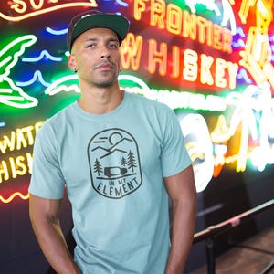 May include: A light blue t-shirt with a black graphic design featuring a mountain scene, trees, and a camper van, with the text "IN MY ELEMENT". The person is wearing a green and black baseball cap.