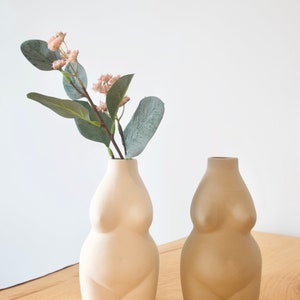 May include: Two ceramic vases shaped like the human body. The vase on the left is white and the vase on the right is beige. Both vases are on a wooden surface. A sprig of greenery with pink flowers is in the white vase.