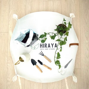 Peut inclure: Un tapis de jardinage blanc avec une plante, des outils de jardinage, un arrosoir et un sac de terre. Le tapis porte l'inscription "HIRAYA POTS AND STANDS".