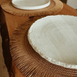 Peut inclure: Deux assiettes en céramique blanche avec une frange tissée brune autour des bords. Les assiettes sont sur une surface en bois.
