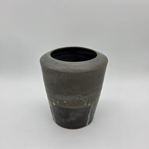 May include: A handcrafted, dark gray ceramic vase with a wide, rounded rim and a cylindrical body. The vase features a textured surface with horizontal bands of varying shades of gray and white accents. The interior is dark.