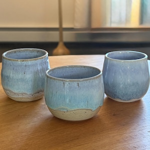 May include: Three handmade ceramic cups with a light blue glaze. The cups have a rounded shape and a textured, speckled appearance. The bottom of each cup has a natural, unglazed band. The cups are displayed on a wooden surface.