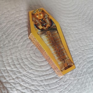 Può includere: Una scatola decorativa a forma di bara gialla con l'immagine di un faraone egizio. La scatola presenta dettagliati motivi geroglifici e una tonalità dorata, che evoca l'antico Egitto.