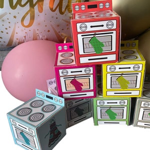 May include: A collection of colorful, miniature oven-shaped boxes. The boxes are red, pink, yellow, green, and blue, each with a printed oven design and a hanging oven mitt. A pink balloon and a gold balloon are in the background.