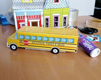 Peut inclure: Un bus scolaire jaune en papier avec le texte "SCHOOL PARTY BUS" et le numéro "36" est à côté d'une maison en papier avec une façade rouge et verte. La maison a un toit gris et une cheminée. Le bus a des roues noires.