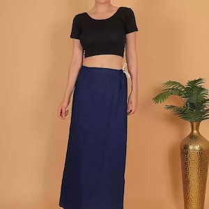 May include: A woman wearing a black crop top and a long, navy blue wrap skirt. The skirt is tied at the waist with a white string. A gold vase with green plants is in the background.