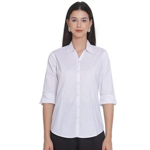 Può includere: Una camicia bianca abbottonata con colletto classico, abbottonatura frontale e maniche a tre quarti. La camicia è sartoriale con cuciture verticali per una vestibilità strutturata ed è abbinata a pantaloni scuri.