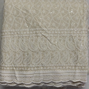 Puede incluir: Tela color crema con intrincada bordado blanco. El diseño presenta un patrón geométrico, motivos florales y formas de cachemira. La tela está doblada, mostrando la textura detallada y el diseño.