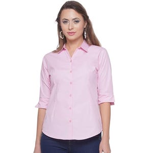 Pode incluir: Uma camisa rosa claro com botões e mangas três quartos. A camisa tem uma gola clássica e um corte ajustado. O tecido parece liso e de cor sólida. A camisa é usada com jeans azuis.
