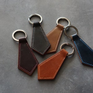 May include: A collection of leather keychains in various colors, including brown, black, and navy blue. Each keychain features a geometric shape with a silver key ring. The stitching is a contrasting color, adding a decorative element.