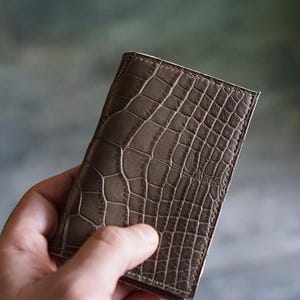 Pode incluir: Uma carteira retangular marrom com textura de pele de crocodilo. A carteira é segurada em uma mão, mostrando o padrão detalhado e as variações de cor. O couro parece ser um marrom suave com mudanças de tom sutis.
