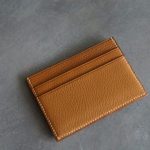 May include: A tan leather card holder with three card slots. The card holder has a textured surface and white stitching around the edges. The background is a solid gray color.