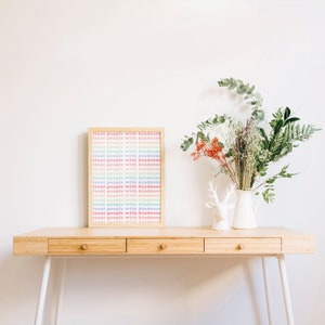 May include: A framed print with the text "treat people with kindness" repeated in rainbow colors on a light wood background. The print is sitting on a light wood desk with a white vase and a deer figurine.