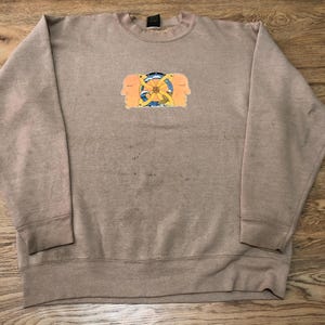 May include: A tan-colored sweatshirt with a crew neck. The front features a graphic of two faces flanking a central design with a yellow flower and blue and yellow accents. The sweatshirt is made of a soft material.