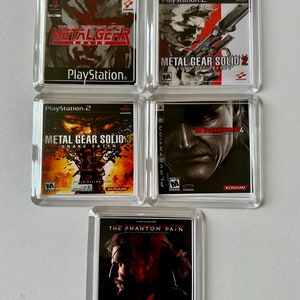 Op de afbeelding: Een verzameling van vier videospelcases voor de Metal Gear Solid-serie. De cases zijn allemaal in heldere kunststof frames met zwart-wit afbeeldingen van de personages en speltitels. De titels omvatten "Metal Gear Solid", "Metal Gear Solid 2: Sons of Liberty", "Metal Gear Solid 3: Snake Eater" en "Metal Gear Solid V: The Phantom Pain".
