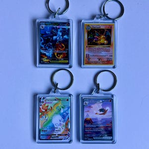 May include: Four clear acrylic keyrings featuring Charizard Pokémon trading cards. The cards display colourful illustrations of Charizard in various poses, with silver key rings attached at the top. The background is a light blue.