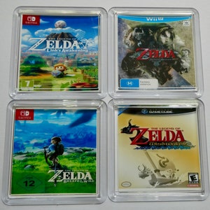 May include: Four different video game cartridges for the Legend of Zelda series. The cartridges are displayed on clear coasters. The games are Link's Awakening, Twilight Princess HD, Breath of the Wild, and Wind Waker.